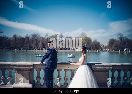 Rückansicht der jungen eleganten Paar in Brautkleider lehnt Am Zaun am See an sonnigen Tagen Stockfoto