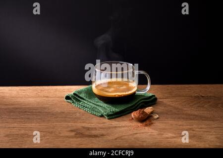 Glas Tasse aromatischen heißen schwarzen Kaffee auf grüner Serviette Platziert mit Löffel gemahlenen Zimt auf Holztisch mit Schwarzer Hintergrund Stockfoto