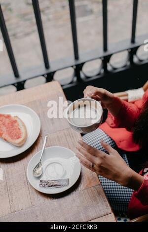 Von oben beschnittene unkenntliche Frau, die an einem kleinen Holztisch saß, beim Frühstück und beim Kaffeetrinken im Café Stockfoto