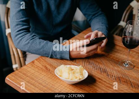 Schneiden Sie den Mann mit dem Telefon und überprüfen Sie die Nachricht, während Sie sitzen Kleiner Holztisch mit einem Glas Wein und Snacks Café Stockfoto