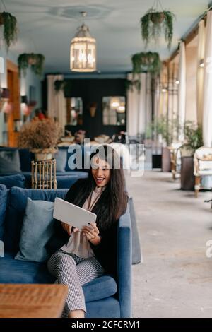 Glückliche junge Frau, die ein Tablet benutzt, während sie auf einer bequemen Couch in der Lobby sitzt Stockfoto