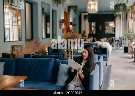 Glückliche junge Frau, die ein Tablet benutzt, während sie auf einer bequemen Couch in der Lobby sitzt Stockfoto