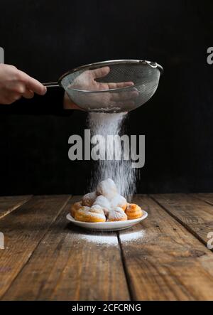 Ernte unkenntlich kochen sieben Zuckerpulver auf schmackhafte Kekse auf Teller auf Holztisch vor schwarzem Hintergrund Stockfoto