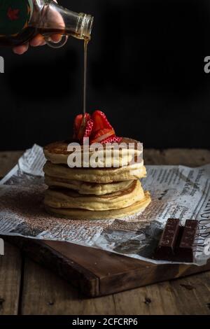 Crop unkenntlich Person Gießen Ahornsirup auf appetitlich leckeren Stapel Von Pfannkuchen mit reifen Erdbeeren auf der Oberseite und Schokolade serviert Auf Papier Stockfoto