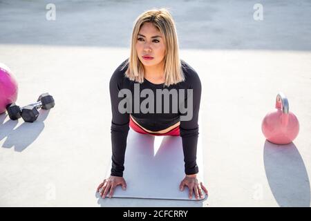 Von oben blonde junge Frau in Sportkleidung, die wegschaut, während sie sich auf geraden Händen lehnt, die auf einer Matte auf dem Betondach des Gebäudes liegen Stockfoto