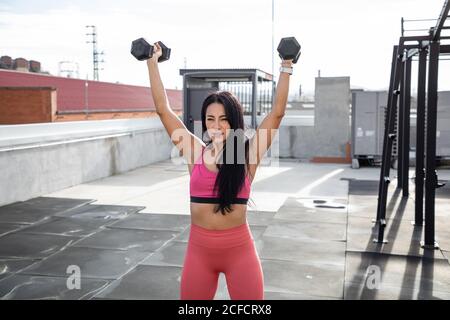 Schwarzhaarige weibliche Sportlerin in rosa aktiv tragen in der Hocke Position mit Hanteln in den Händen auf Dach des Gebäudes suchen Weg Stockfoto