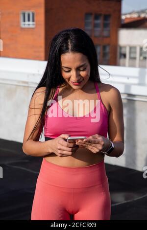 Junge zufrieden Brünette in stilvollen rosa BH und Leggings lächelnd Während des Surfens Handy während des Trainings auf dem Dach des Gebäudes Stockfoto