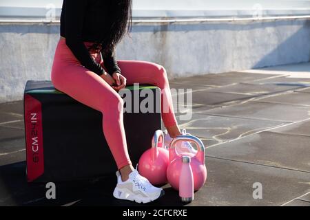 Crop Beine von weiblichen Athleten in stilvollen rosa Leggings und Weiße Turnschuhe sitzen auf Box mit Hanteln und Flasche auf Dach des Gebäudes Stockfoto