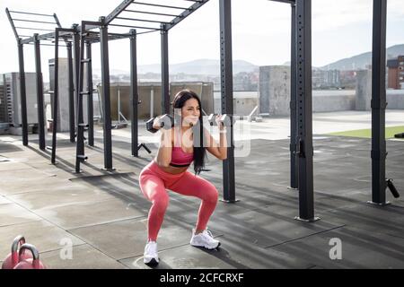 Schwarzhaarige weibliche Sportlerin in rosa aktiv tragen in der Hocke Position mit Hanteln in den Händen auf Dach des Gebäudes suchen Weg Stockfoto