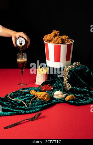 Ernte weibliche Gießen süße Soda aus der Dose in Weinglas neben Luxuriöser Goldschmuck und Portionen gebratener Hähnchendrumsticks und pommes frites in der Zusammensetzung mit Ketchup und Mayonnaise auf grün Samt gegen schwarze Wand Stockfoto