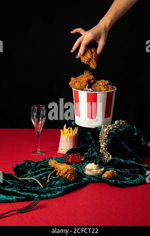 Crop Frau, die gebratene Chicken Nuggets aus dem Eimer auf dem roten Tisch mit goldenem Schmuck in Komposition mit Portionen pommes Frites und Saucen auf grünem Samt nimmt Stockfoto