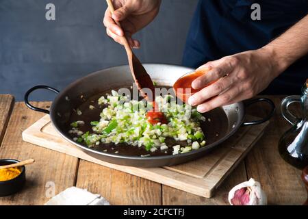 Von oben unkenntlich kochen Zugabe von Tomatenmark mit Holzspatel Zu gehackter grüner Chilischote und Zwiebel auf groß gemischt Metallpfanne während der Zubereitung der Zutaten in der modernen Küche Zu Hause Stockfoto