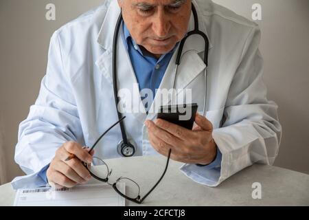 Ältere männliche Hausarzt in medizinischen Kleid sitzen am Tisch Mit verschreibungspflichtigen Notebook in der modernen Klinik und Kommunikation auf dem Smartphone Per Videoanruf mit dem Patienten während einer Coronavirus-Pandemie Stockfoto
