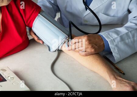 Von oben des Getreidearztes mit Stethoskop, das am Tisch sitzt Im medizinischen Raum und Messung des Blutdrucks mit Tonometer während Arbeiten im Krankenhaus während des Coronavirus-Ausbruchs Stockfoto