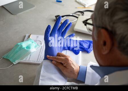 Von oben der Ernte männlichen Hausarzt setzen auf Einweg Chirurgische Handschuhe während der Arbeit in der Klinik während covid 19 Pandemie Stockfoto