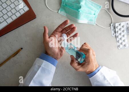 Draufsicht des männlichen Therapeuten im medizinischen Kleid sitzend an Tisch im Krankenhaus und desinfizieren Hände mit antiseptischen während der Vorbereitung Zur Behandlung von Patienten während des Coronavirus-Ausbruchs Stockfoto