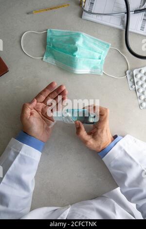 Draufsicht des männlichen Therapeuten im medizinischen Kleid sitzend an Tisch im Krankenhaus und desinfizieren Hände mit antiseptischen während der Vorbereitung Zur Behandlung von Patienten während des Coronavirus-Ausbruchs Stockfoto