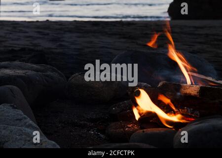 Nahaufnahme des brennenden Lagerfeuers mit großen Steinen am Meer platziert Abends mit Meerwasser im Hintergrund Stockfoto