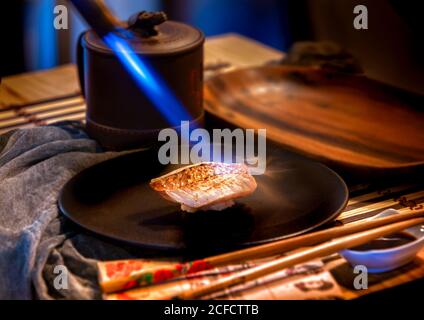 Blaues Licht des Brenners Vorbereitung Stück Fisch auf spezielle Teller und Sauce mit Stöcken auf dem geservierten Tisch Stockfoto