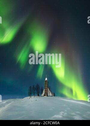 Erstaunliche Landschaft von leuchtenden bunten Nordlichtern in dunklen Himmel Über schneebedecktes Gelände mit kleiner Dorfkirche und Bäumen Island Stockfoto