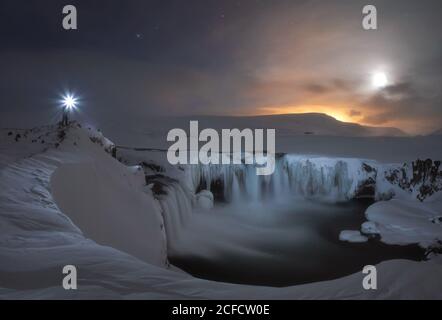 Einsamer unerkennbarer Reisender mit glühender Laterne, die auf einem Felsen in der Nähe steht Kleiner gefrorener See mit Wasserfällen inmitten schneebedecktem Wüstengelände Sonnenuntergang in Island Stockfoto