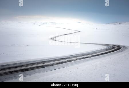 Leere kurvige Landstraße, die durch schneebedecktes Wüstengelände führt Kalter Wintertag mit blauem Himmel in Island Stockfoto