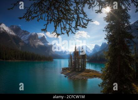 Erstaunliche kanadische Landschaft mit winzigen Insel mit Fichten in Schöner ruhiger See mit türkisfarbenem Wasser, umgeben von majestätischen Felsen Berge mit Schnee bedeckt, umrahmt von Ästen von Nadelbäumen An sonnigen Tagen Stockfoto