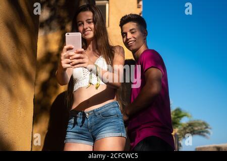 Von unten freudige junge Frau, die Selfie auf dem Smartphone nimmt Mit einem glücklicher ethnischer Mann, der sich am sonnigen Tag am Strand entspannen kann Stockfoto