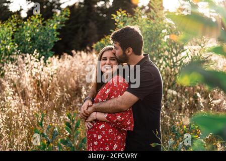 Pensive Mann umarmt zärtlich lächelnd schwangere Frau auf Hintergrund Malerischer grüner Park an sonnigen Tagen Stockfoto