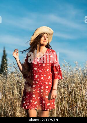 Attraktive lächelnde Frau in Strohhut und rotem Kleid, die auf dem wilden Feld auf dem Hintergrund des blauen Himmels steht Stockfoto