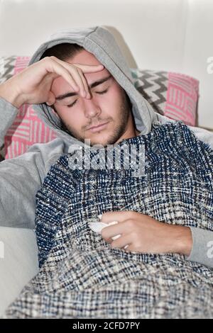 Nahaufnahme eines jungen Mannes, der einen Kapuzenpullover mit einer Serviette trägt und unter einer Decke auf einem Sofa ruht. Krankheit und Gesundheitskonzept. Stockfoto