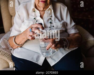 Gealterte lockige Frau im weißen Hemd, die Pillen mit Wasser aus der Flasche trinkt, die auf einem Sessel in der Wohnung sitzt Stockfoto