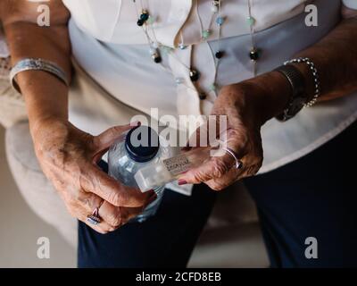 Gealterte lockige Frau im weißen Hemd, die Pillen mit Wasser aus der Flasche trinkt, die auf einem Sessel in der Wohnung sitzt Stockfoto