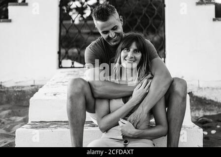 Schwarz-weiß fröhlicher erwachsener Mann und Frau, die lächeln und umarmen, während sie auf Treppen vor dem Gebäude sitzen Stockfoto