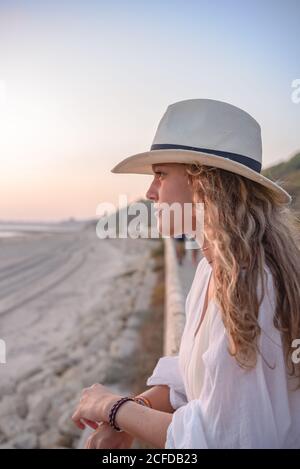Seitenansicht einer anmutigen Frau mit langen lockigen Haaren im Hut, die sich am Zaun lehnt und auf das Meer blickt Stockfoto