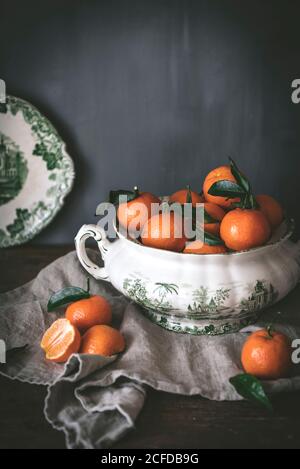 Anspruchsvolles Stillleben mit reifen orangen Mandarinen mit grünen Blättern In tiefweißer Schale auf Tisch auf grauem Hintergrund Stockfoto