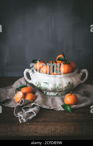 Anspruchsvolles Stillleben mit reifen orangen Mandarinen mit grünen Blättern In tiefweißer Schale auf Tisch auf grauem Hintergrund Stockfoto