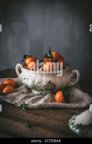 Anspruchsvolles Stillleben mit reifen orangen Mandarinen mit grünen Blättern In tiefweißer Schale auf Tisch auf grauem Hintergrund Stockfoto