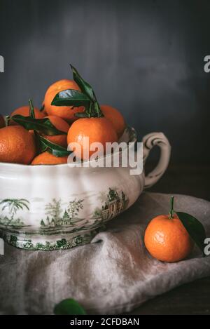 Anspruchsvolles Stillleben mit reifen orangen Mandarinen mit grünen Blättern In tiefweißer Schale auf Tisch auf grauem Hintergrund Stockfoto