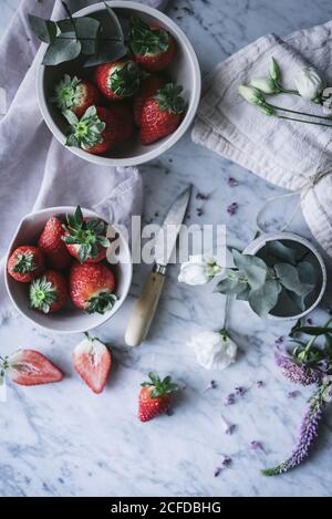Schalen mit reifen Erdbeeren und schönen Blumen auf weiß platziert Marmorplatte in der Nähe des Messers Stockfoto