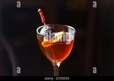 Roter Alkoholcocktail manhattan garniert mit Kirsch- und Orangenschale auf Stick vor dunklem Hintergrund Stockfoto