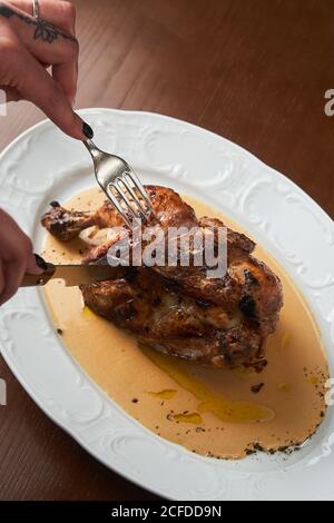 Von oben anonyme Frau hält Besteck und Schneiden gegrilltes Huhn Auf Platte auf Tisch Stockfoto