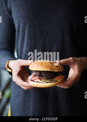 Crop anonyme Person in schwarz tragen halten klassische Burger mit Cutlet und Gemüse mit Käse, während die Fast-Food-Industrie Stockfoto