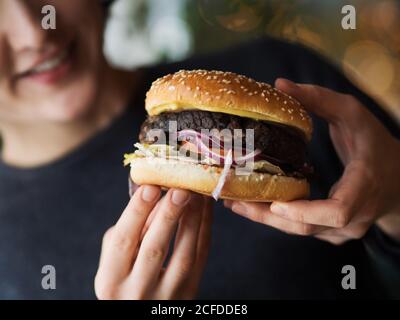 Crop anonyme Person in schwarz tragen halten klassische Burger mit Cutlet und Gemüse mit Käse, während die Fast-Food-Industrie Stockfoto