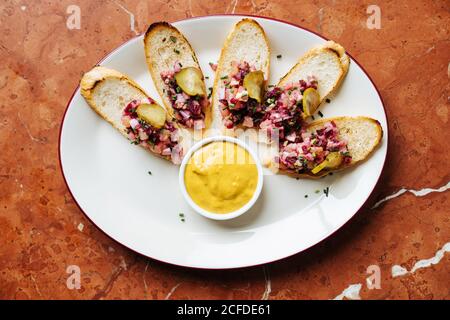 Draufsicht auf geröstete Brotscheiben mit buntem Salat Und abgerundete Stücke von gesalzenen Gurken auf ovalem weißen Teller Mit gelber Sauce Stockfoto