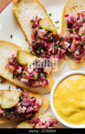 Draufsicht auf geröstete Brotscheiben mit buntem Salat Und abgerundete Stücke von gesalzenen Gurken auf ovalem weißen Teller Mit gelber Sauce Stockfoto