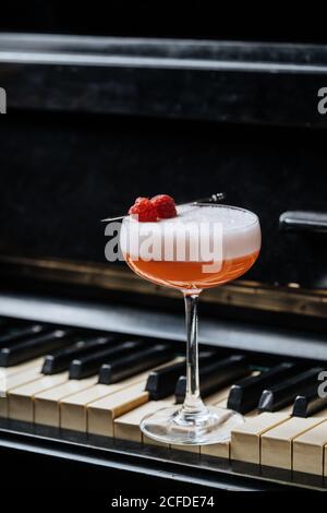 Roter Alkoholcocktail in stilvollem Glas mit weißem Schaumstoff verziert Mit frischer Himbeere auf Klaviertasten im Restaurant Stockfoto