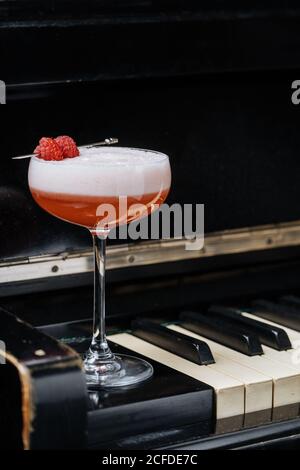 Roter Alkoholcocktail in stilvollem Glas mit weißem Schaumstoff verziert Mit frischer Himbeere auf Klaviertasten im Restaurant Stockfoto