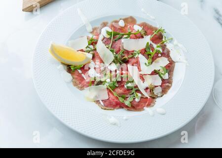 Von oben von Scheiben rotes Rindfleisch auf Teller mit Stücken Von frischer Zitrone mit weißer Sauce Käse und Rucola verziert Stockfoto