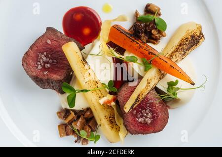 Blick von oben auf köstliche Gourmet geröstete Fleischsteaks mit gegrilltem Gemüse garniert mit frischen Kräutern und Sauce auf weiß serviert Platte Stockfoto